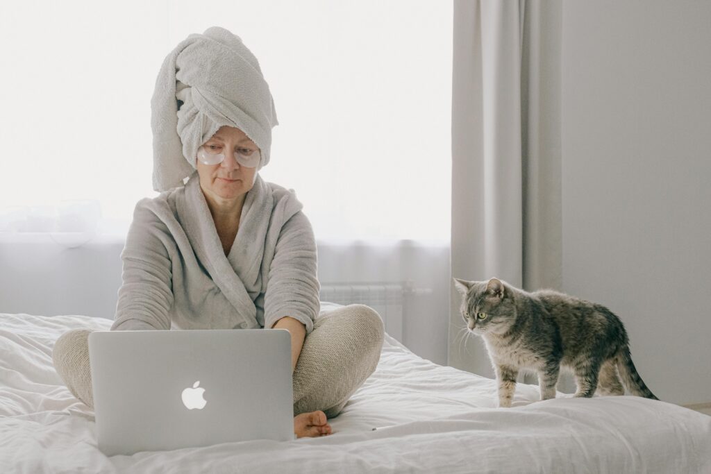 A woman in a robe uses a laptop while a cat sits beside her in a cozy bedroom setting.